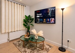 Een woonkamer met een flatscreen-tv waarop een Netflix-interface te zien is, een potplant, een glazen salontafel met decoratieve objecten, een vloerlamp en een radiator.