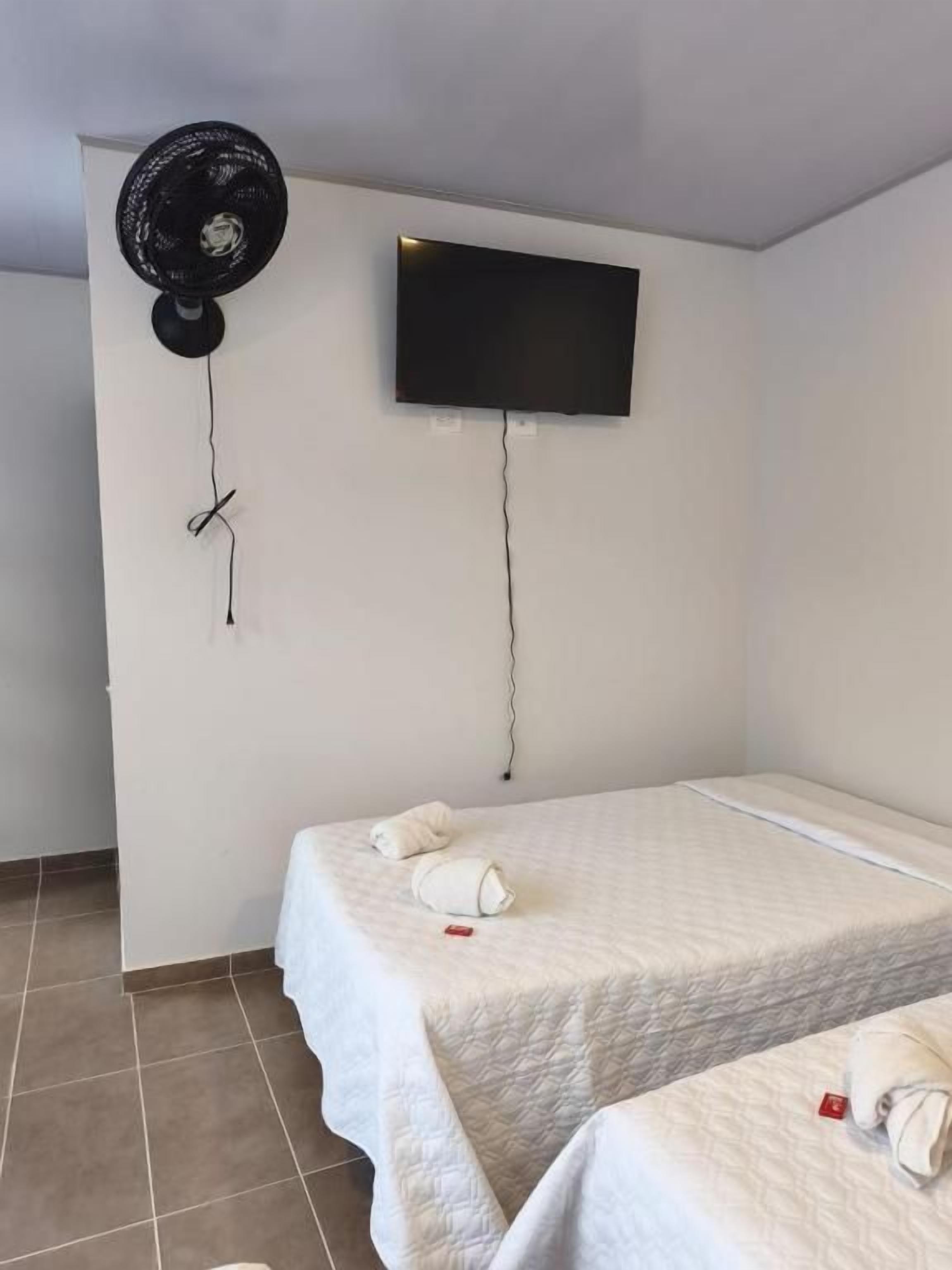 A room with two beds, a flat-screen TV mounted on the wall, and a standing fan.