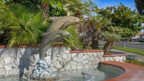 Une fontaine ornée de deux sculptures de dauphins, située dans un espace paysager agrémenté de palmiers et d’un mur de pierre.