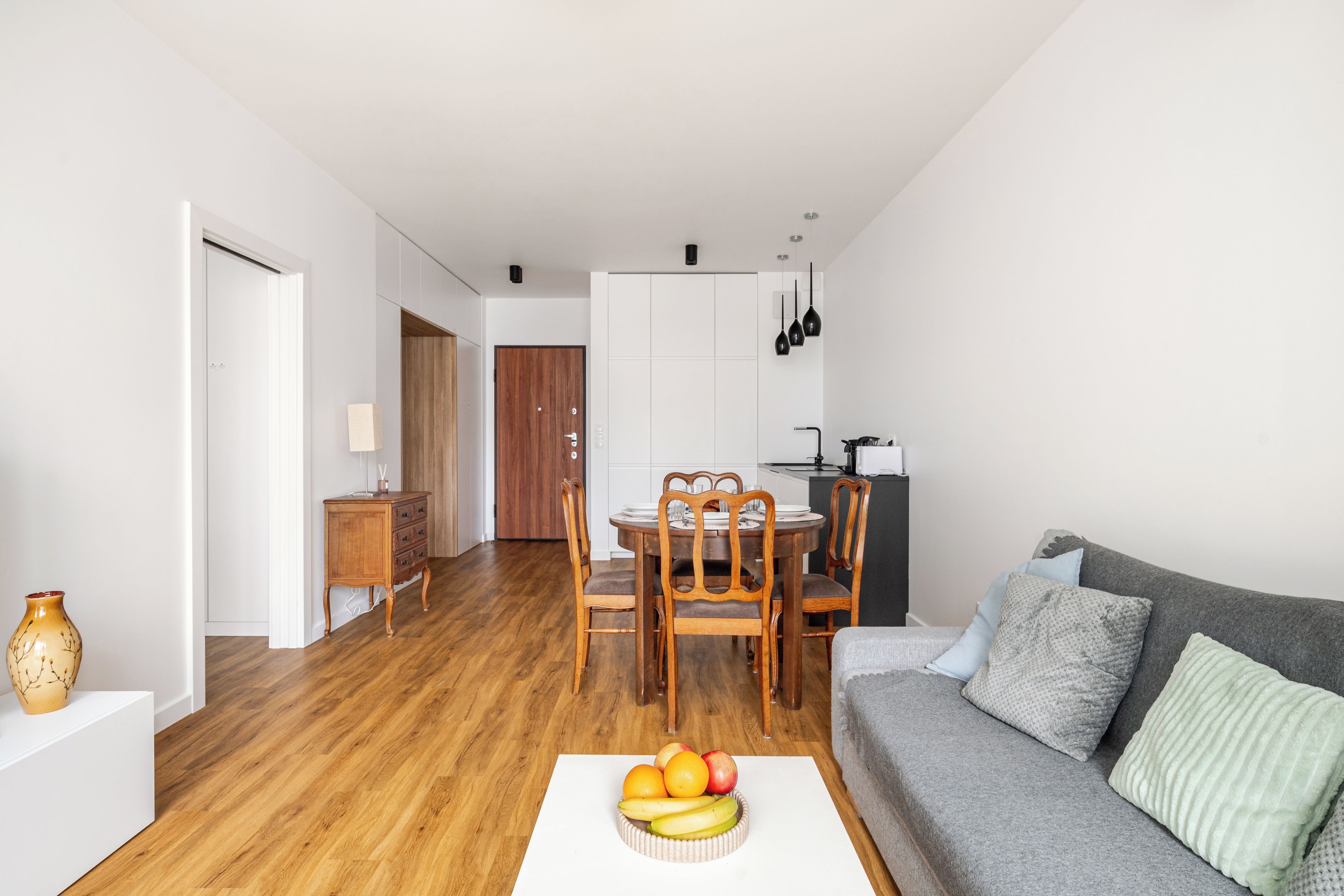 Ein modernes Wohnzimmer mit einem Holzesstisch, einem grauen Sofa und einer Obstschale auf einem weißen Couchtisch.