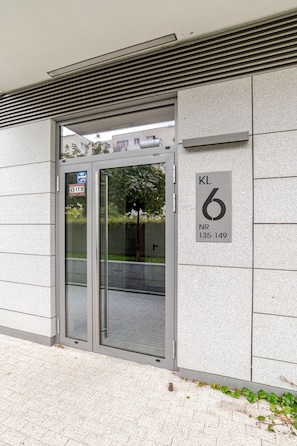 Entrada de um edifício com porta de vidro, placa com 'KL 6' e 'NR 135-149', e aviso de segurança.