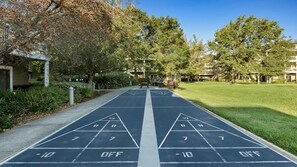 Ein Shuffleboardfeld mit einem klaren Weg zu einem Gebäude, umgeben von Grünflächen und Bäumen.