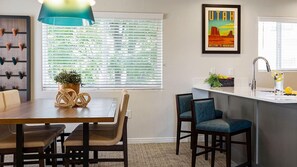 A modern kitchen with a dining table, chairs, a hanging light fixture, a window with blinds, a framed welcome sign, and a kitchen island with a sink and fruit bowl.