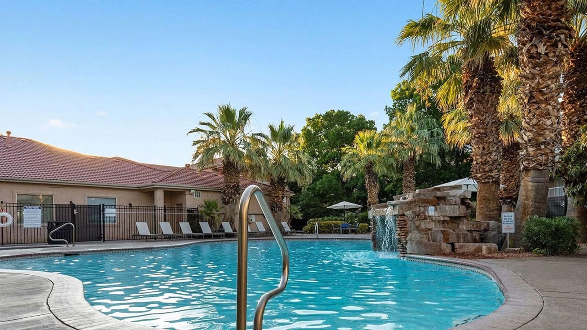 A swimming pool with a metal ladder, lounge chairs, palm trees, and a building with a tiled roof.