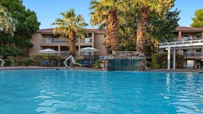 A swimming pool with a waterfall feature, surrounded by palm trees and a building with balconies.