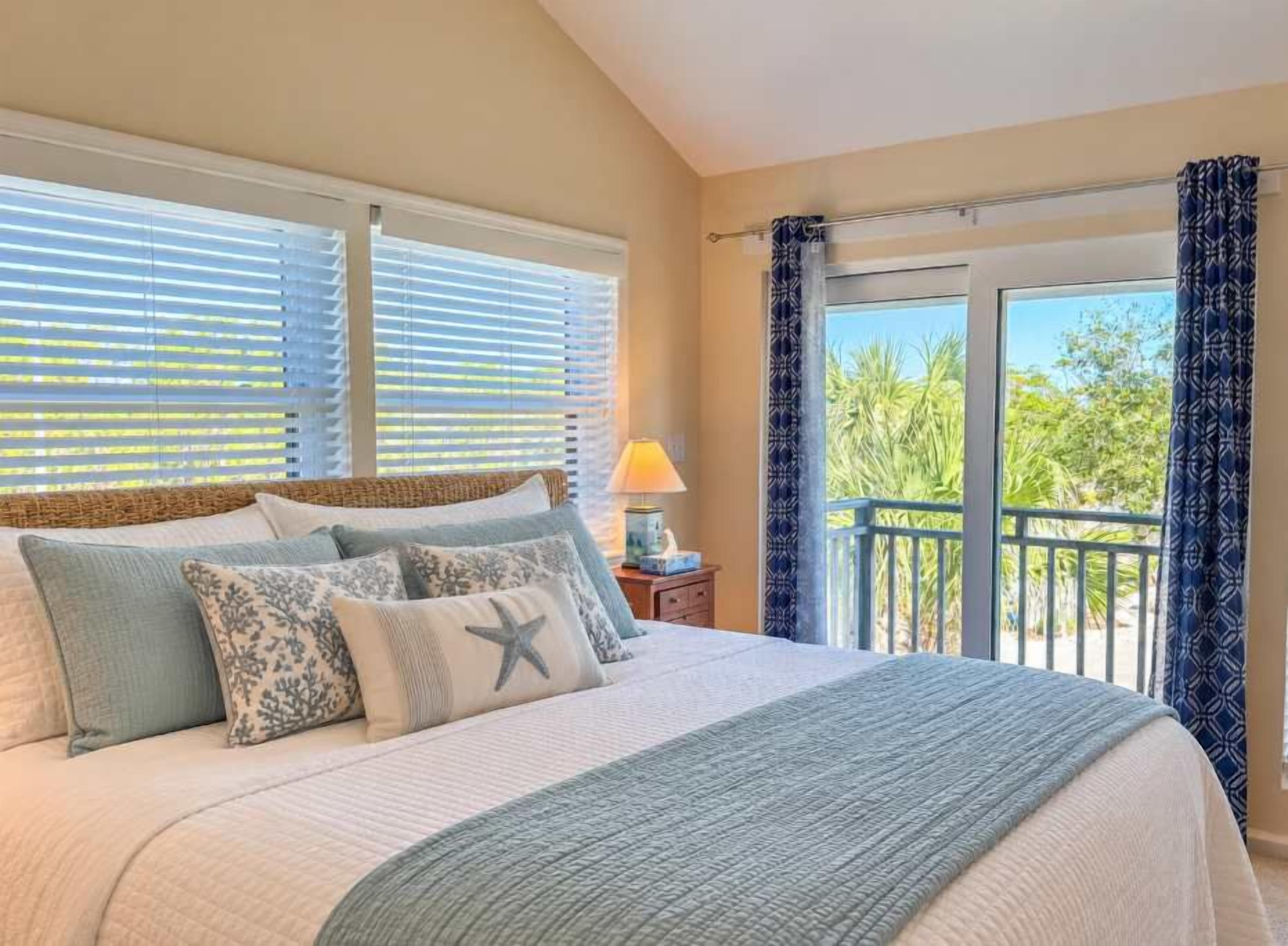 A bedroom with a bed, pillows, a nightstand, a lamp, and a view of the outdoors through a balcony.