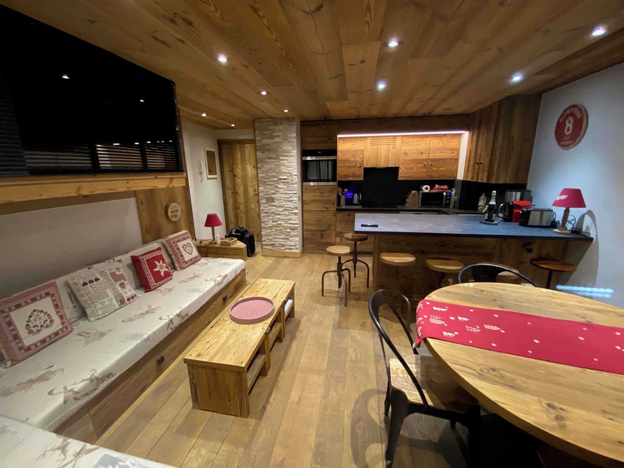 A cozy interior with a wooden ceiling, a stone accent wall, a kitchen area with modern appliances, a dining table with chairs, and a sofa with patterned cushions.