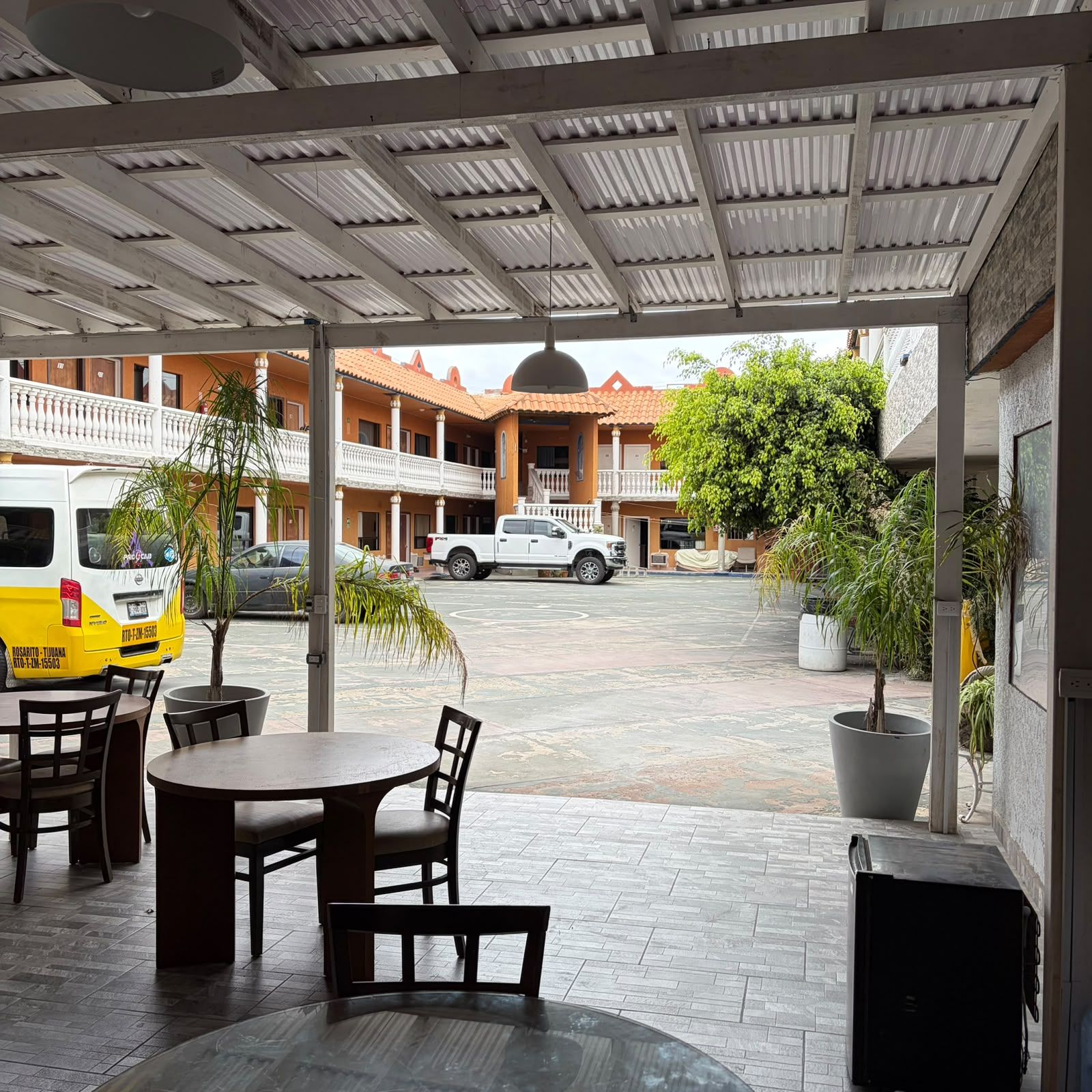 A covered outdoor seating area with tables and chairs, a van, and a building with balconies in the background.