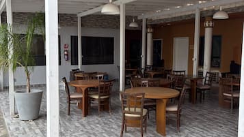 An outdoor cafe area with multiple wooden tables and chairs, a large potted plant, and a fire extinguisher on the wall.