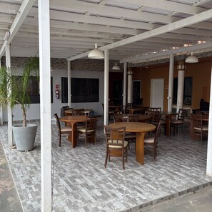 Un área de café al aire libre con varias mesas y sillas de madera, una planta grande en maceta y un extintor en la pared.