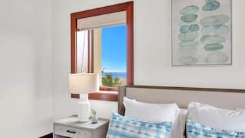 A hotel room with a bed, a nightstand, a lamp, a window with a view of the ocean, and a framed artwork on the wall.
