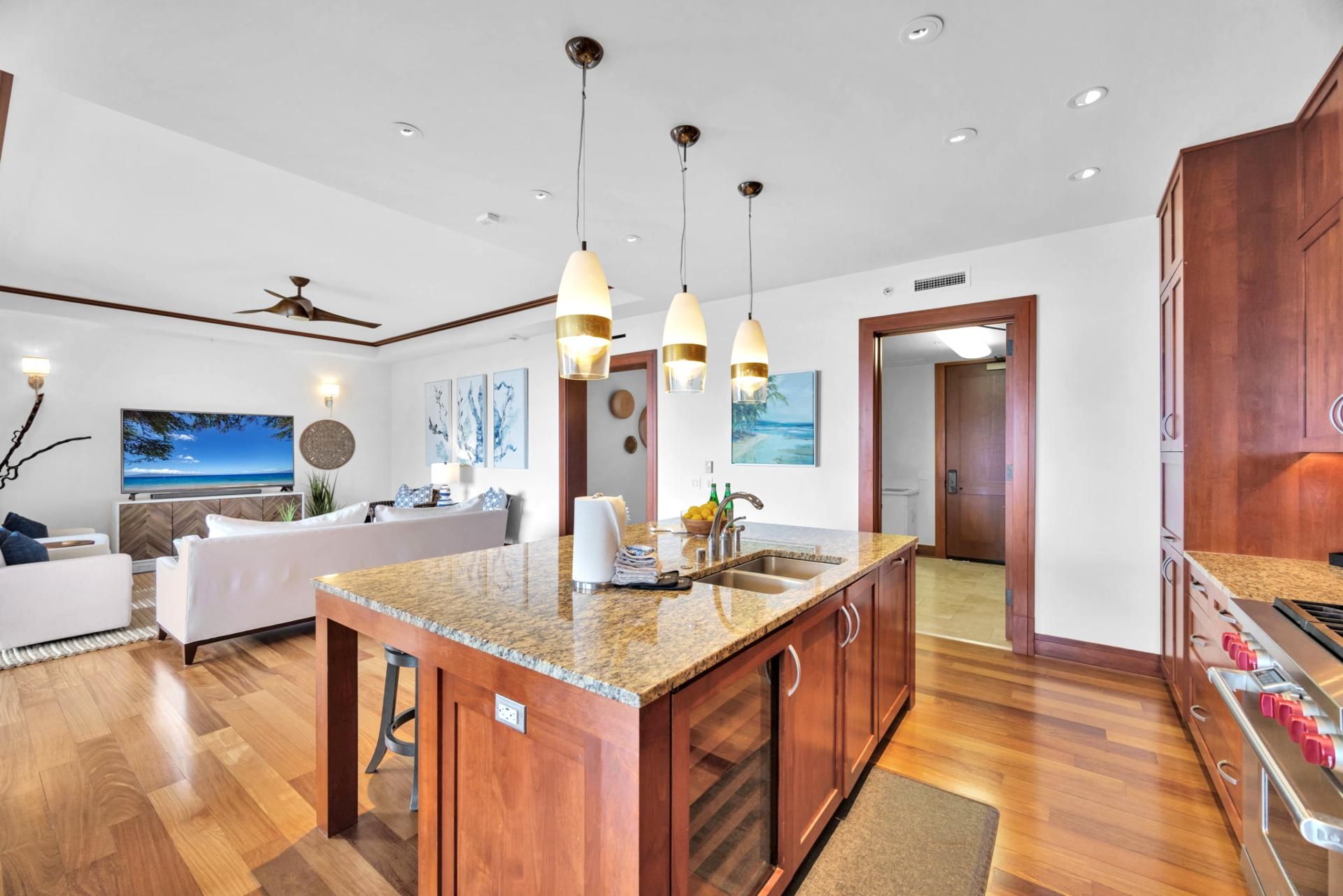 A modern kitchen with granite countertops, wooden cabinets, and stainless steel appliances. There is a dining area with a white table and chairs, and a living room with a sofa and a TV.
