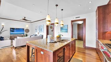 A modern kitchen with granite countertops, wooden cabinets, and stainless steel appliances. There is a dining area with a white table and chairs, and a living room with a sofa and a TV.