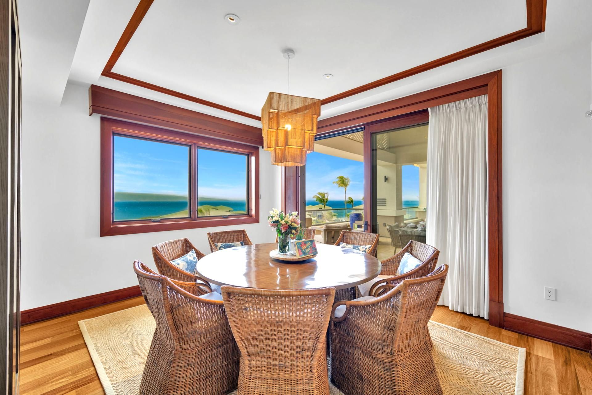 Un coin repas avec une table ronde, des chaises en osier, un vase de fleurs et une vue sur l’océan grâce à de grandes fenêtres.