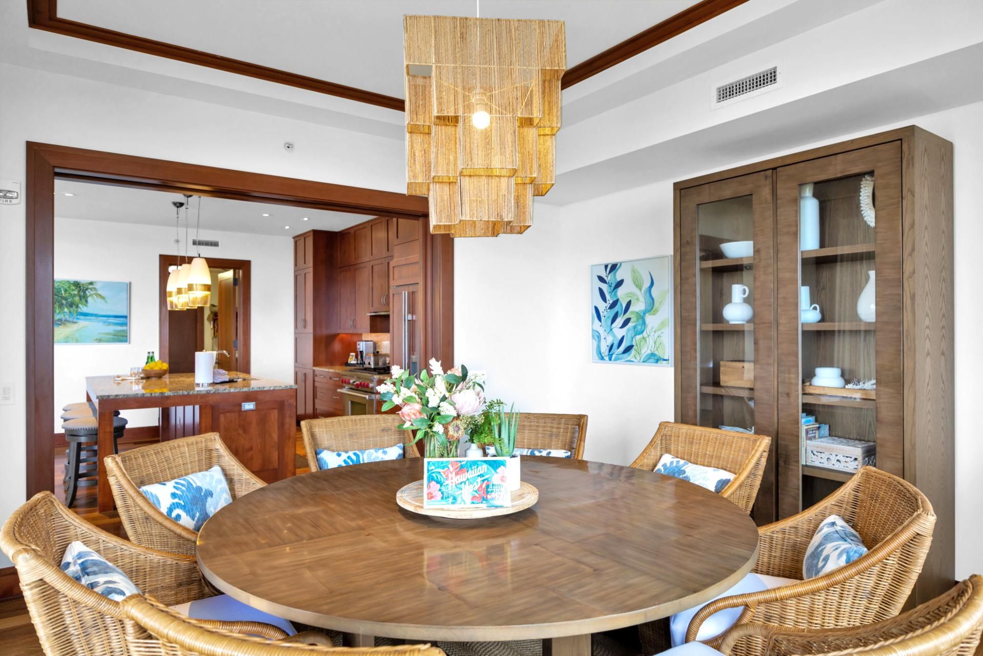 A round wooden dining table surrounded by wicker chairs with blue and white cushions, a wooden cabinet with glass doors, and a hanging light fixture.