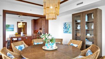 A round wooden dining table surrounded by wicker chairs with blue and white cushions, a wooden cabinet with glass doors, and a hanging light fixture.