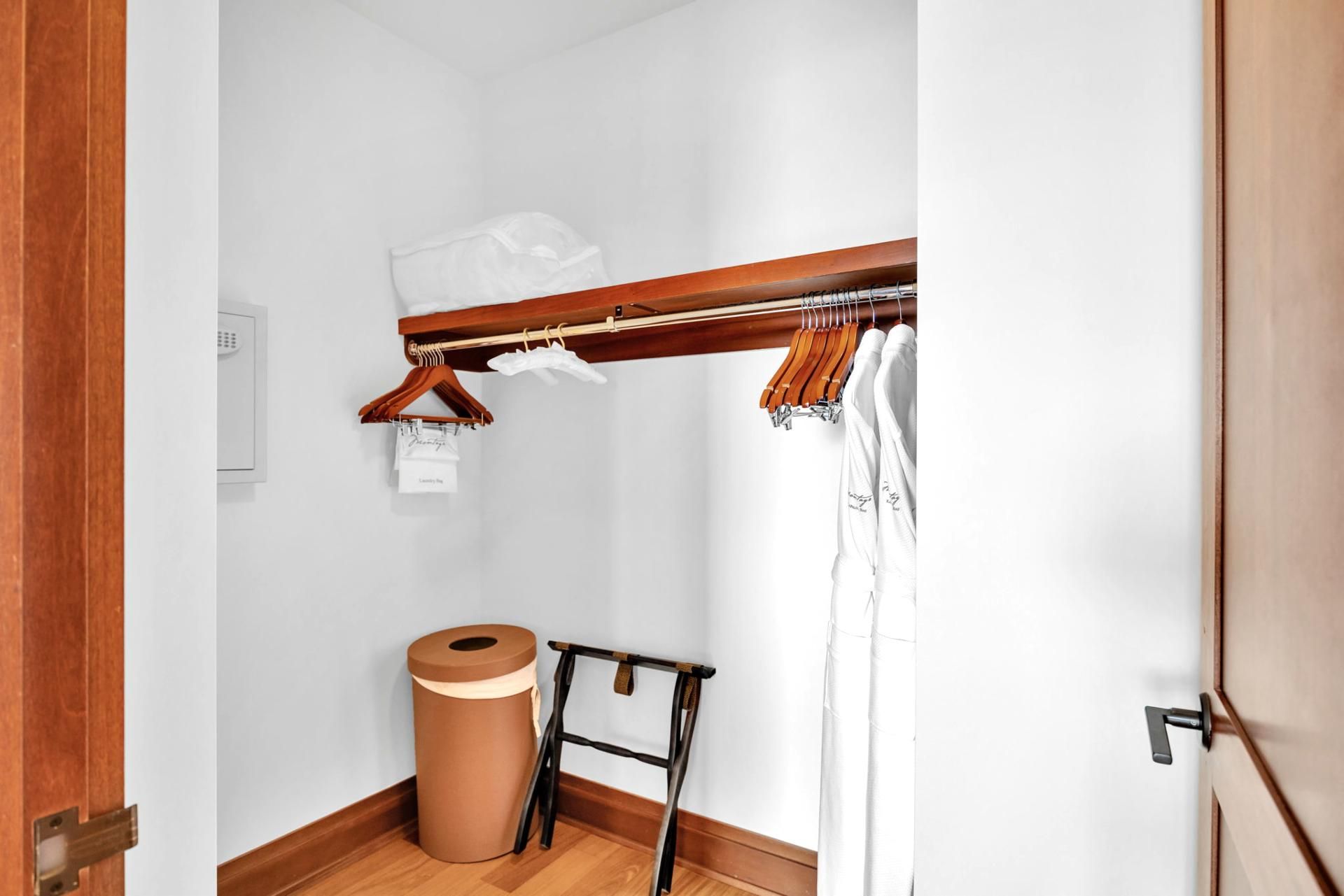 A small, narrow room with a wooden door, a white wall, a wooden floor, a wooden clothes rack, a white towel, a brown trash bin, and a black ladder.