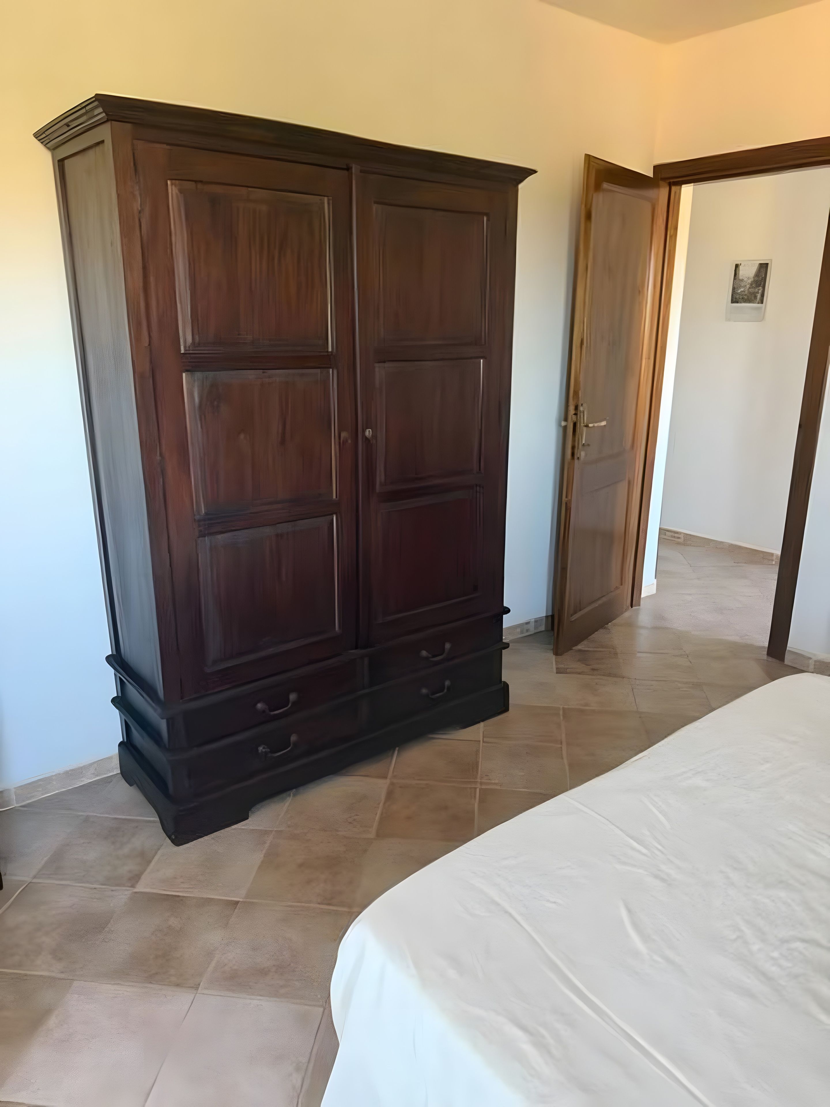 Ein Schlafzimmer mit einem großen, dunklen Holzschrank, einem Bett mit weißer Bettwäsche und einem gefliesten Boden.