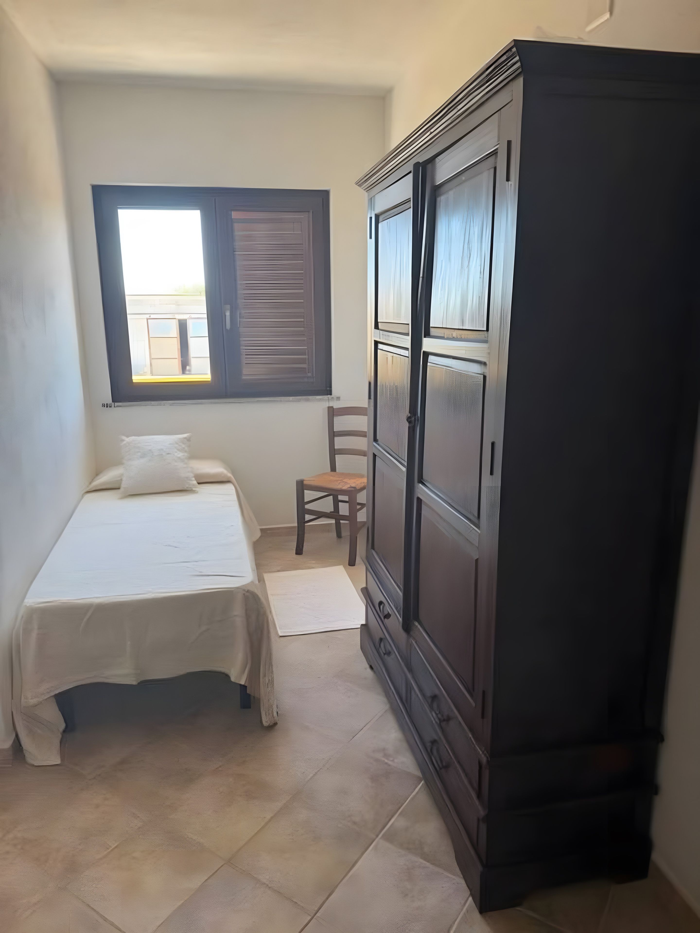 A bedroom with a bed, a wooden chair, a wardrobe, and a window with shutters.