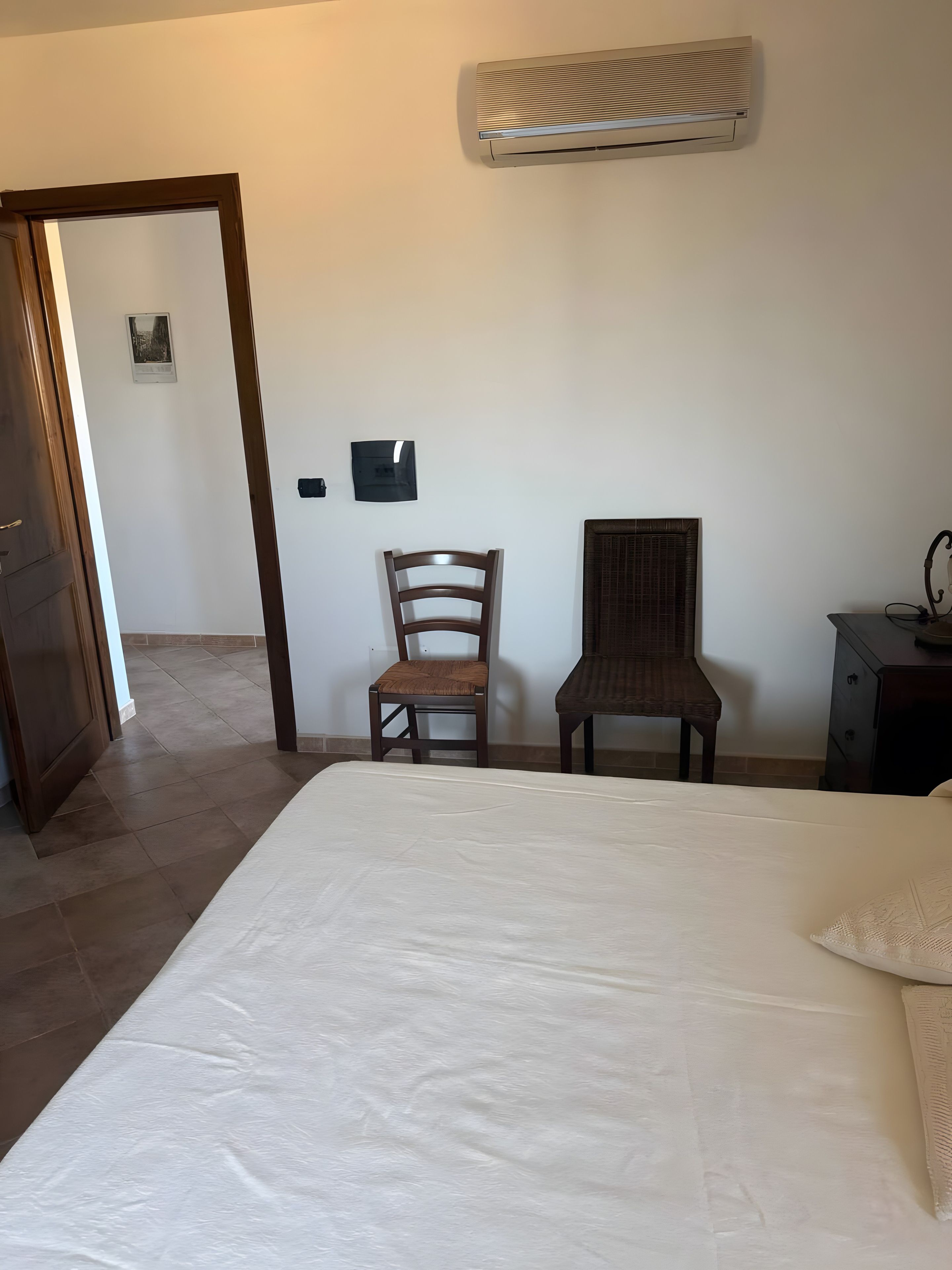 Ein Schlafzimmer mit einem Bett, zwei Sesseln, einem Nachttisch, einer Klimaanlage und einer Tür zu einem anderen Raum.