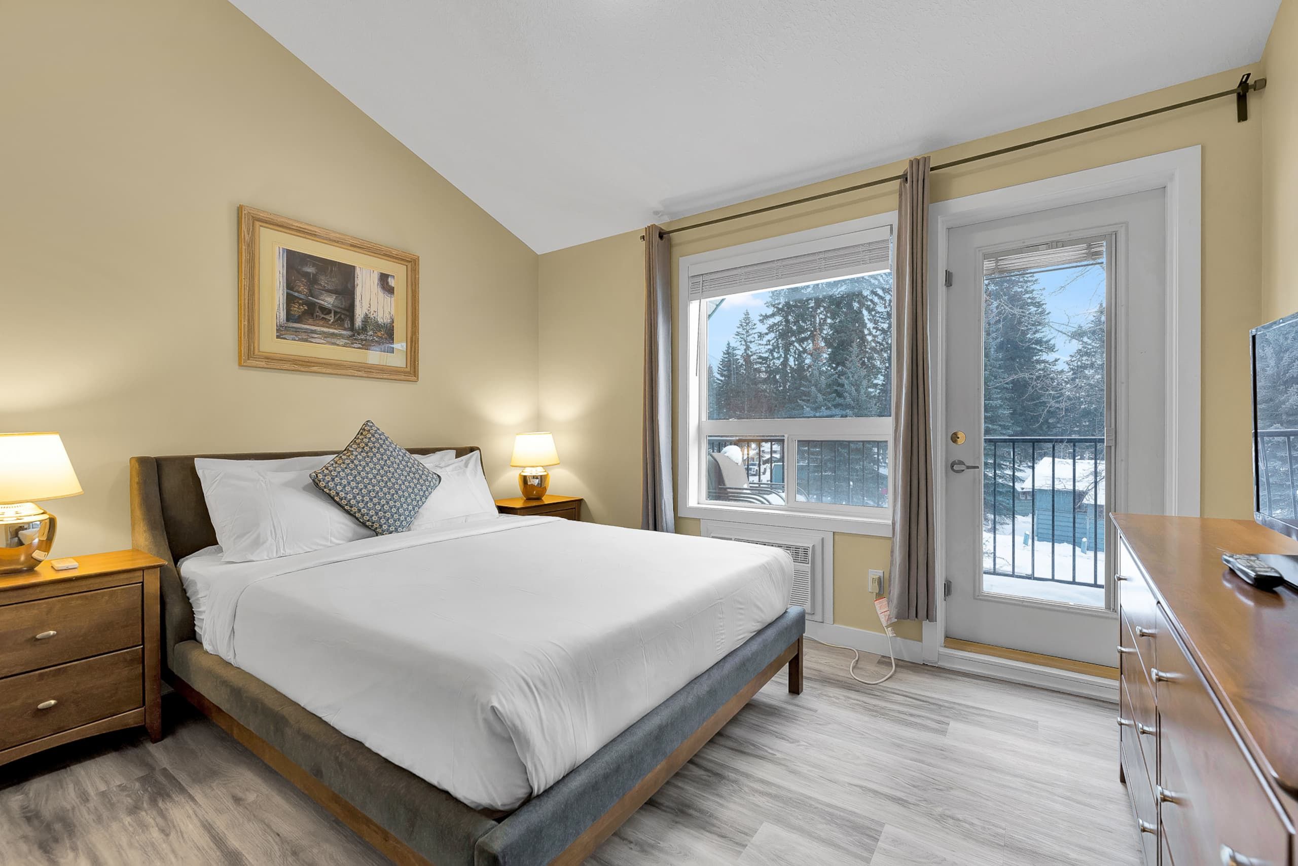 Une chambre à coucher comprenant un lit, des tables de chevet, une commode, une fenêtre donnant sur des arbres et une porte.