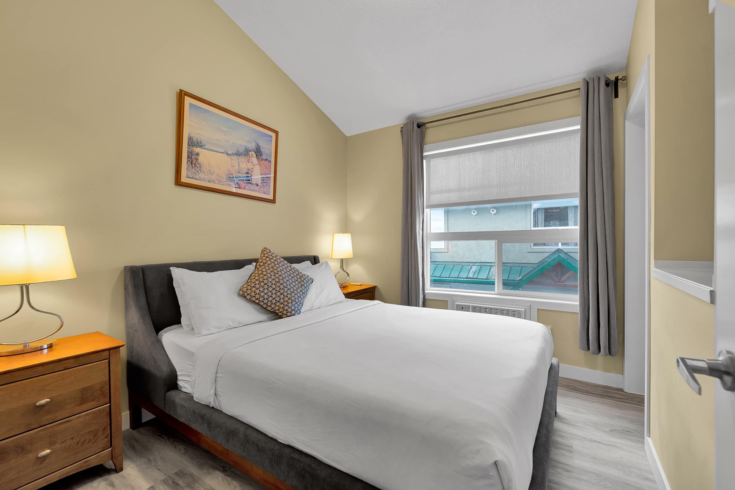 A hotel room with a bed, two bedside lamps, a nightstand, a painting on the wall, and a window with curtains.