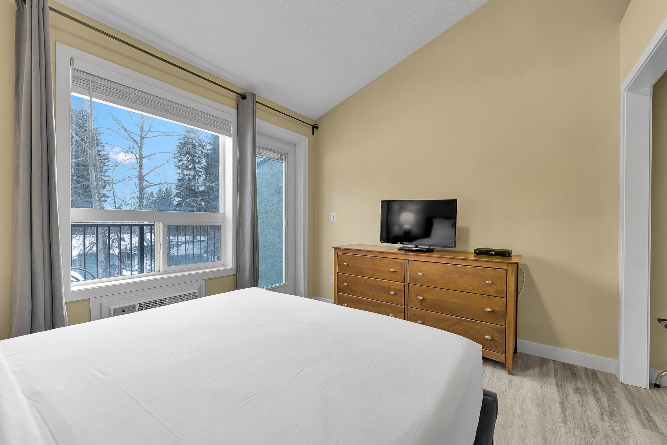 A hotel room with a bed, a dresser, a television, and a window with a view of trees.