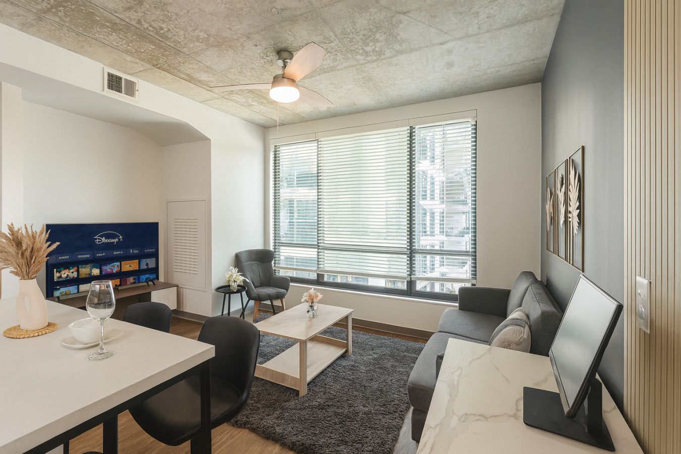 Een moderne woonkamer met een bank, salontafel en stoelen, een flatscreen-tv en een groot raam met jaloezieën.