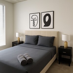 Een netjes opgemaakt bed met twee kussens, een paar opgerolde handdoeken en twee nachtkastjes met lampen.