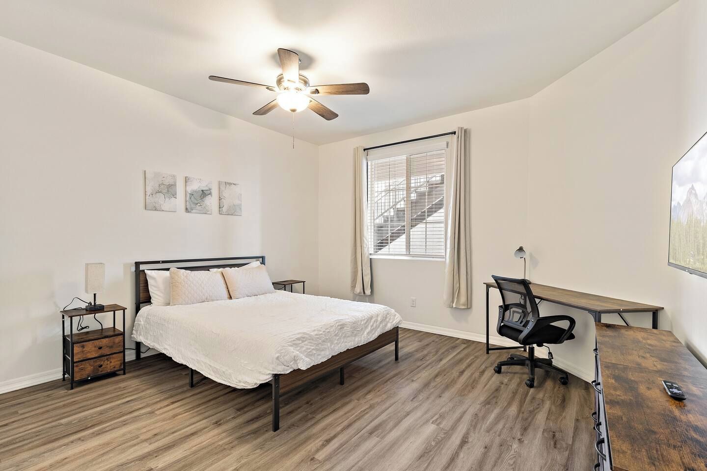 Une chambre à coucher comprenant un lit, un bureau, une chaise, un ventilateur de plafond et une fenêtre avec des stores.
