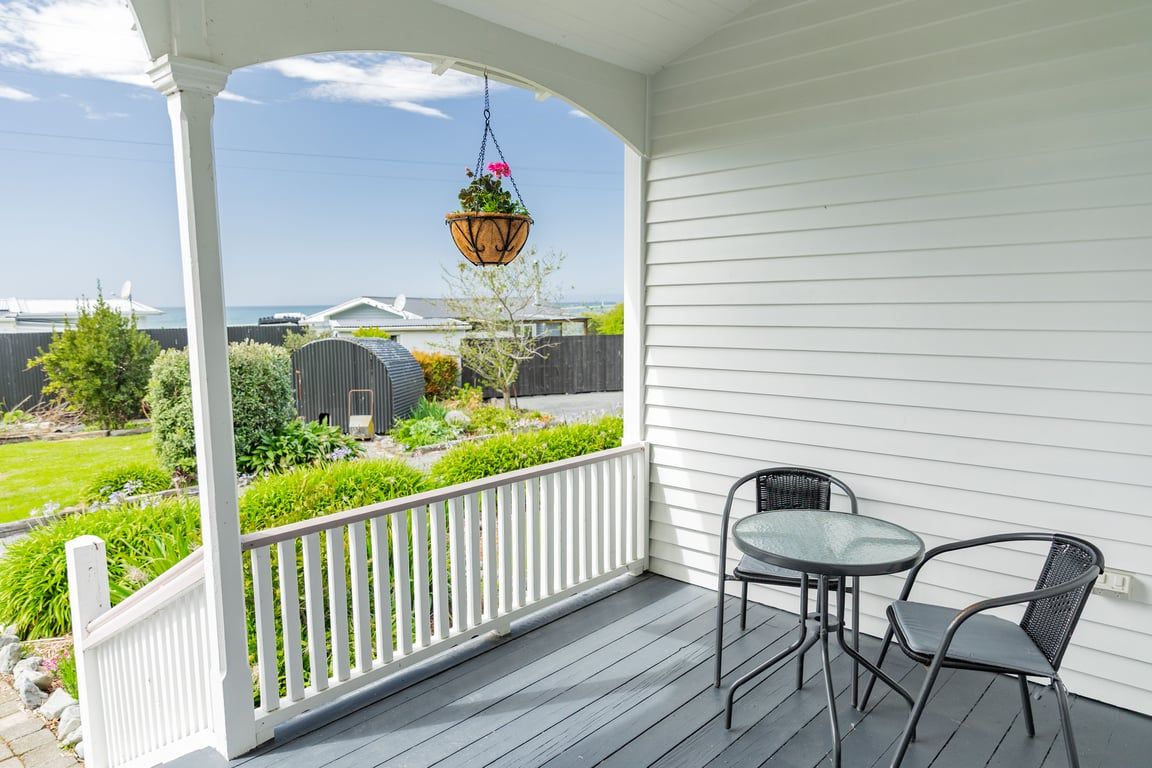 En overdækket veranda med gelænder, en hængende blomsterkasse og et bord med to stole.