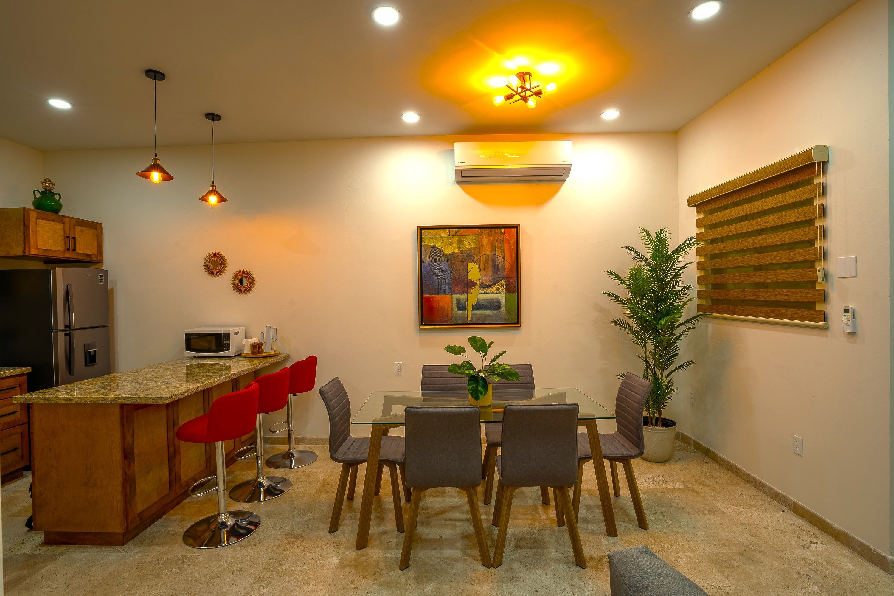 A modern kitchen with a central island, bar stools, a dining table with chairs, and a ceiling light fixture.