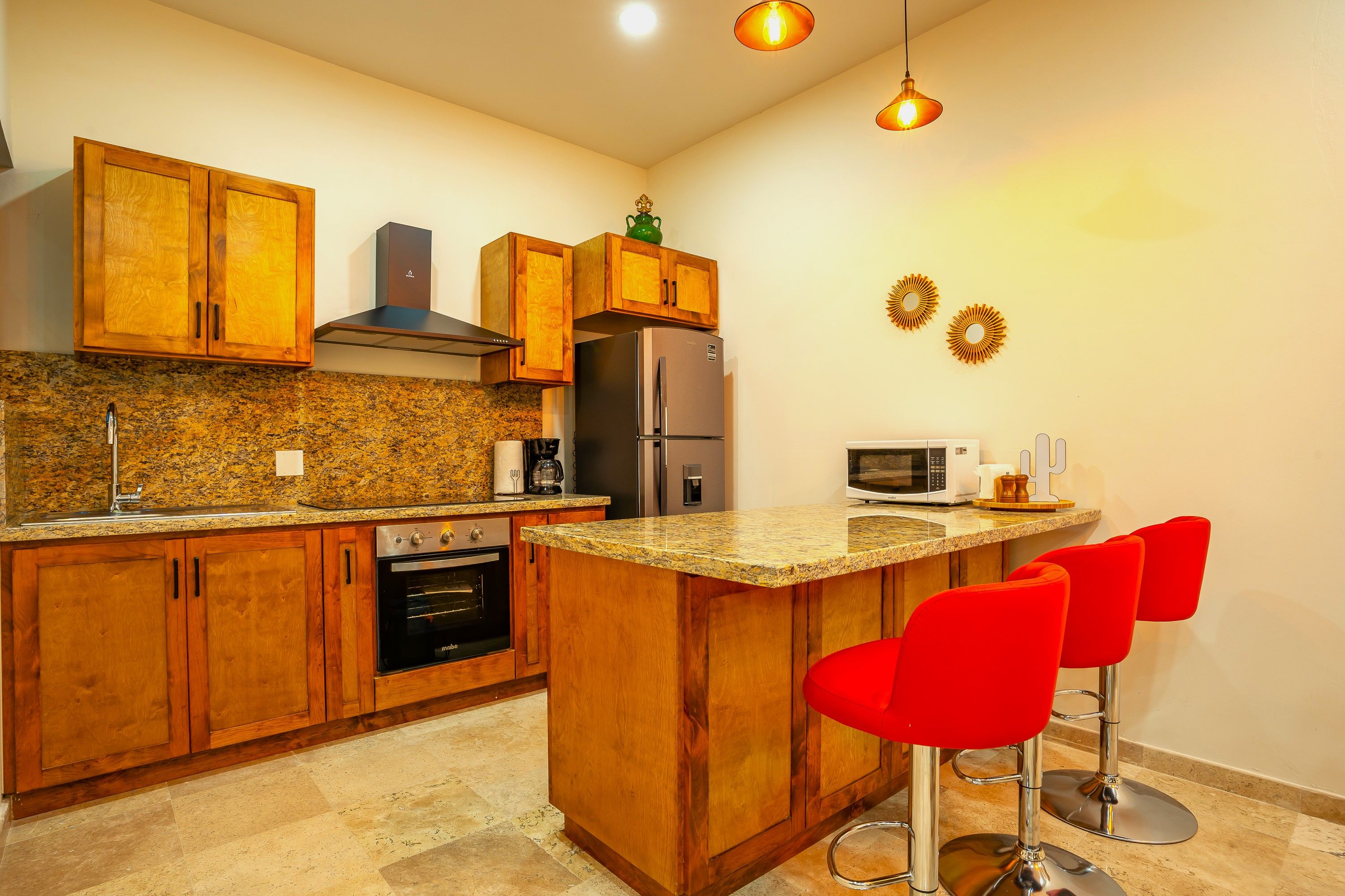 A kitchen with wooden cabinets, a stainless steel refrigerator, a microwave, and a built-in oven. There are two red bar stools and a granite countertop.