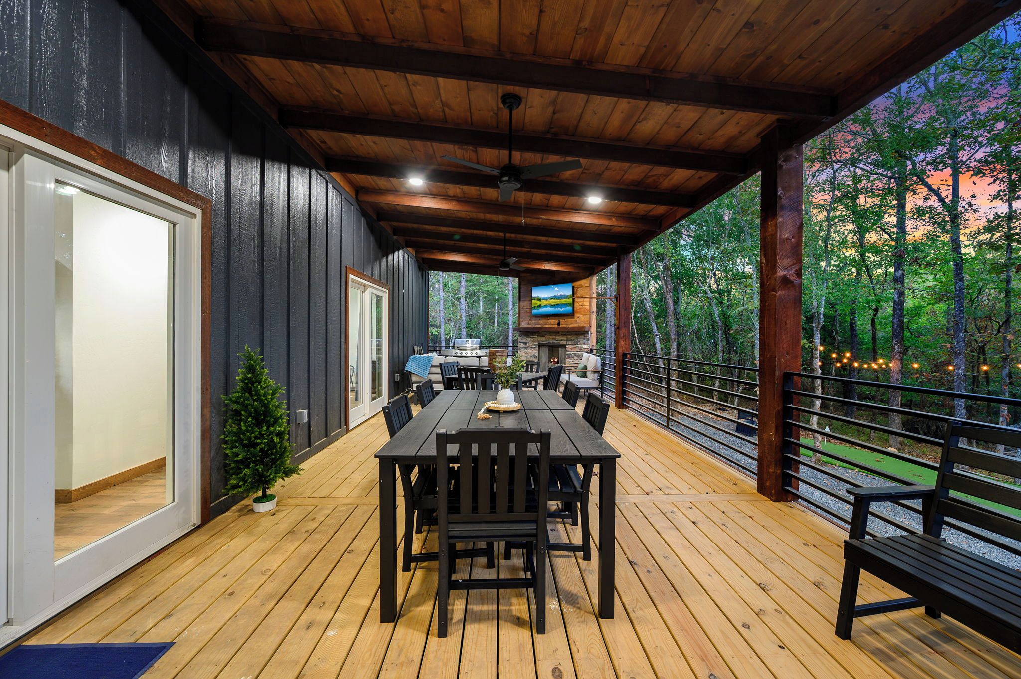 An outdoor covered patio with a dining table and chairs, a television mounted on the wall, and a view of the surrounding forest.