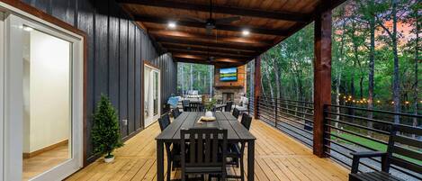An outdoor covered patio with a dining table and chairs, a television mounted on the wall, and a view of the surrounding forest.