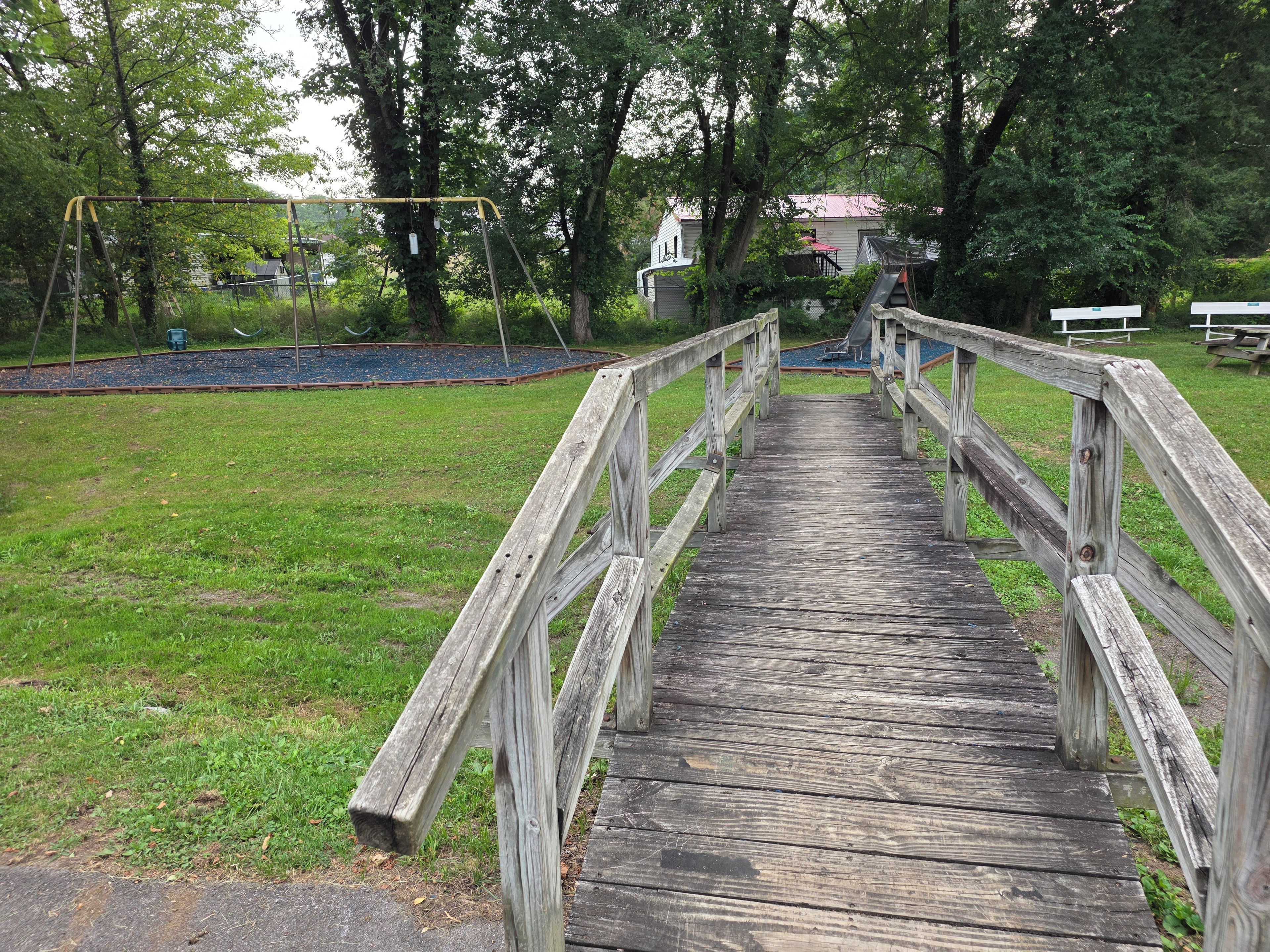 Eine Holzbrücke führt zu einem Spielplatz mit Schaukeln und einer Rutsche.
