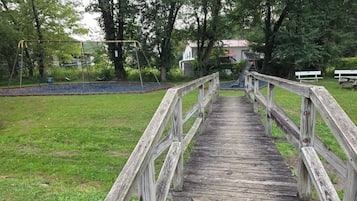 Eine Holzbrücke führt zu einem Spielplatz mit Schaukeln und einer Rutsche.