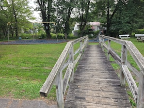 Eine Holzbrücke führt zu einem Spielplatz mit Schaukeln und einer Rutsche.