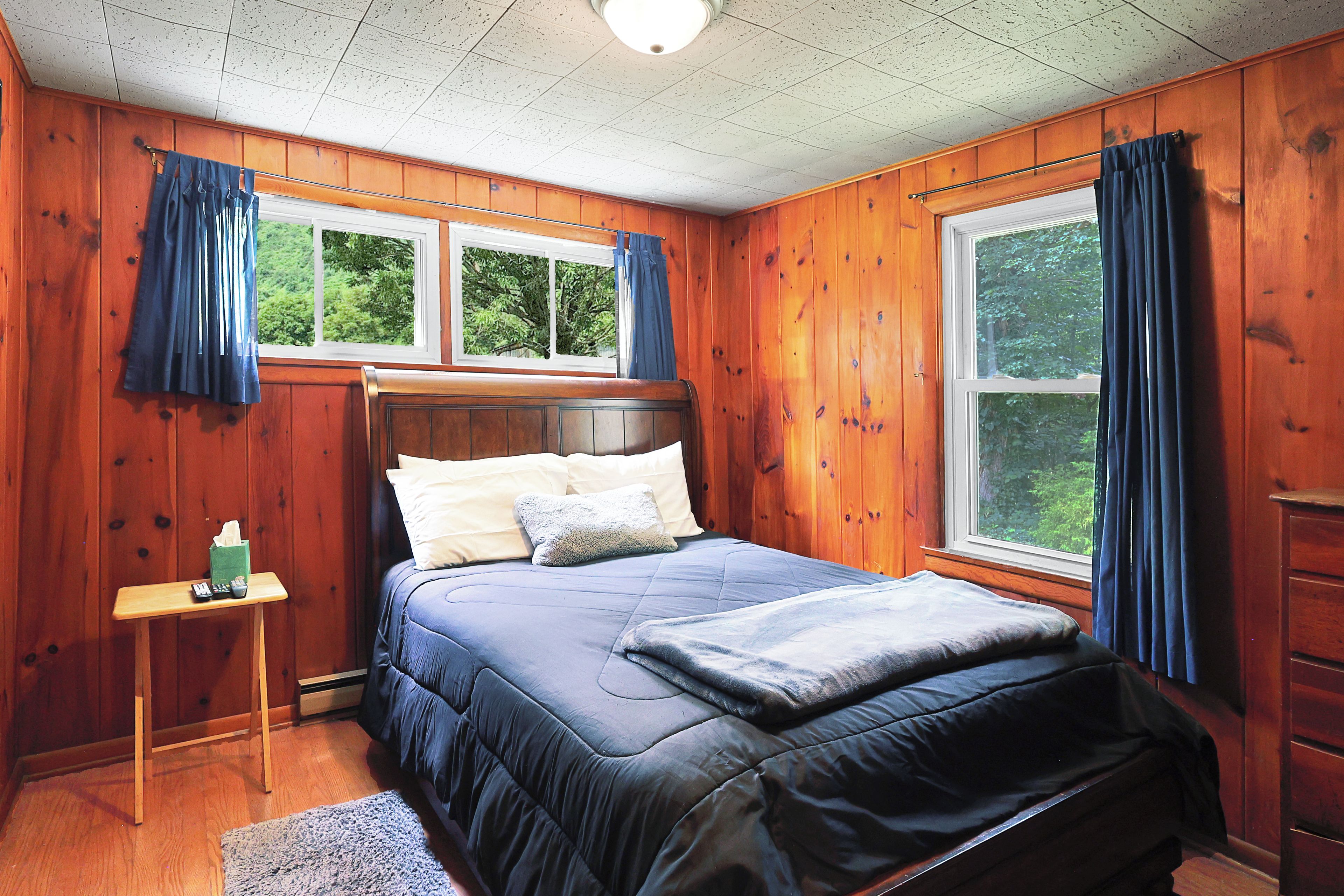 A wooden-paneled bedroom with a bed, two windows, a nightstand, and a dresser.