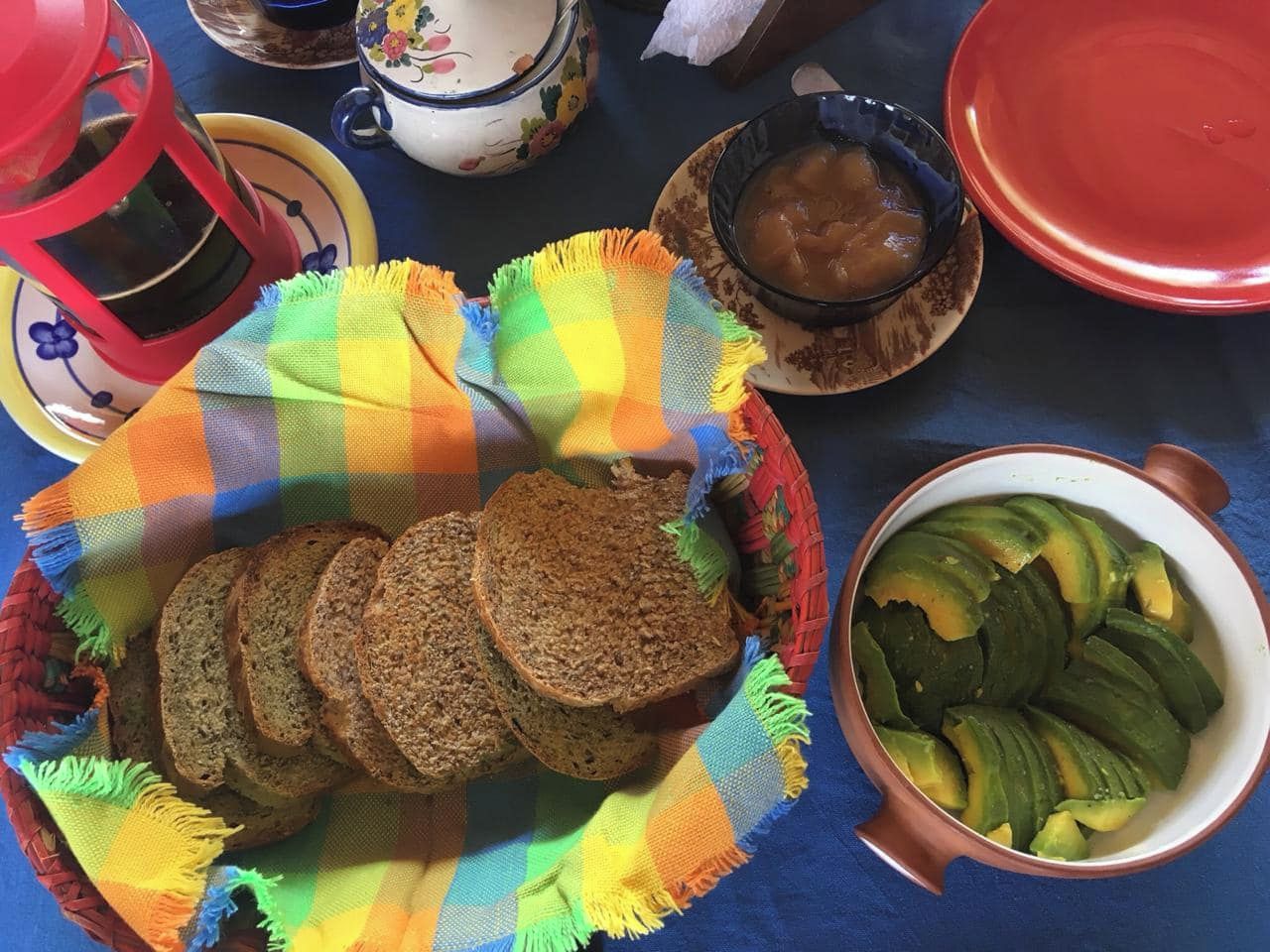 Una mesa con una fuente de pan en rodajas, un tazón de aguacate en rebanadas y una tetera.