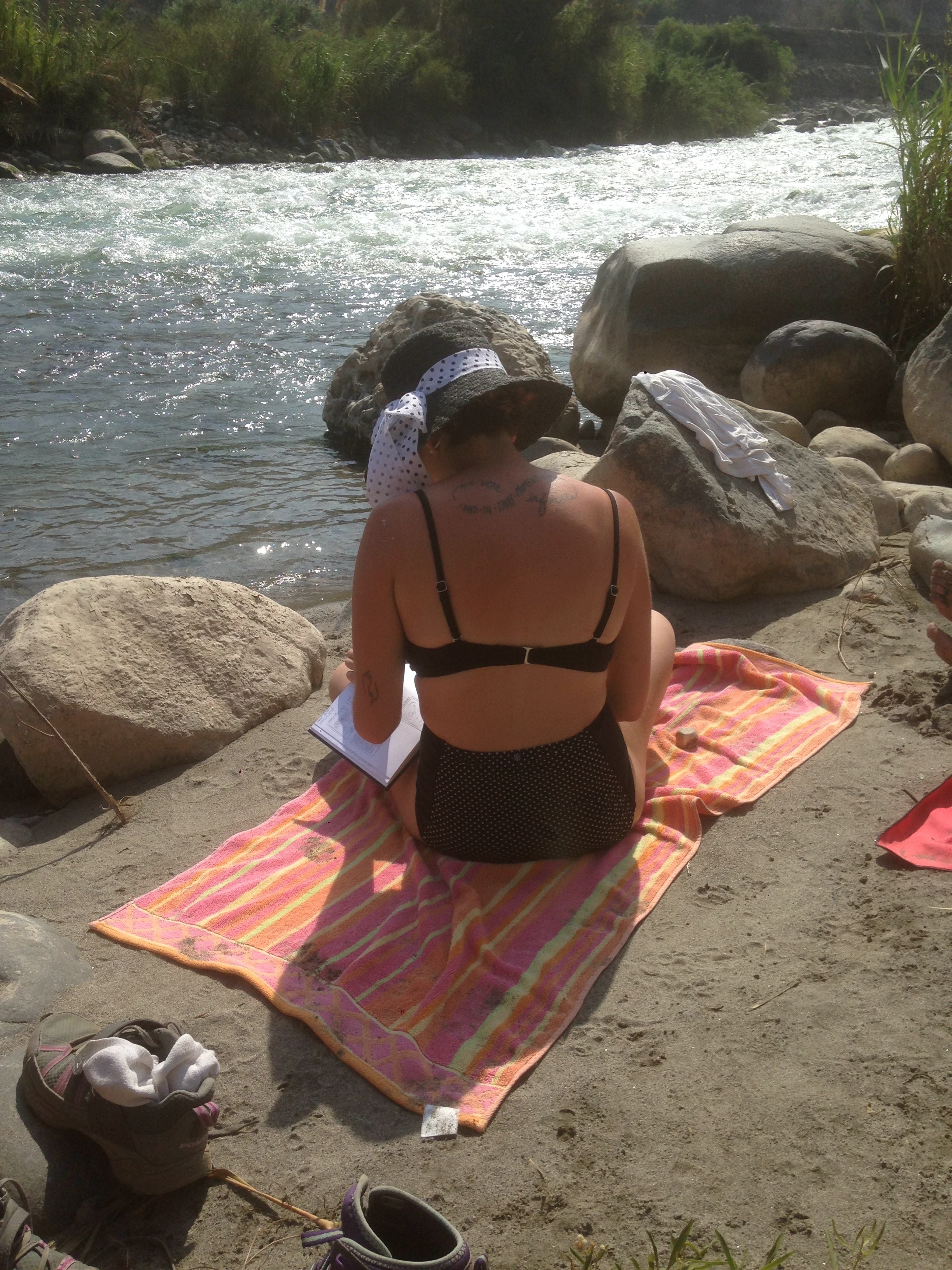 Una persona sentada sobre una toalla en la playa, cerca de un río, con rocas y vegetación al fondo.