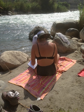A person sitting on a beach towel near a river, with rocks and greenery in the background.