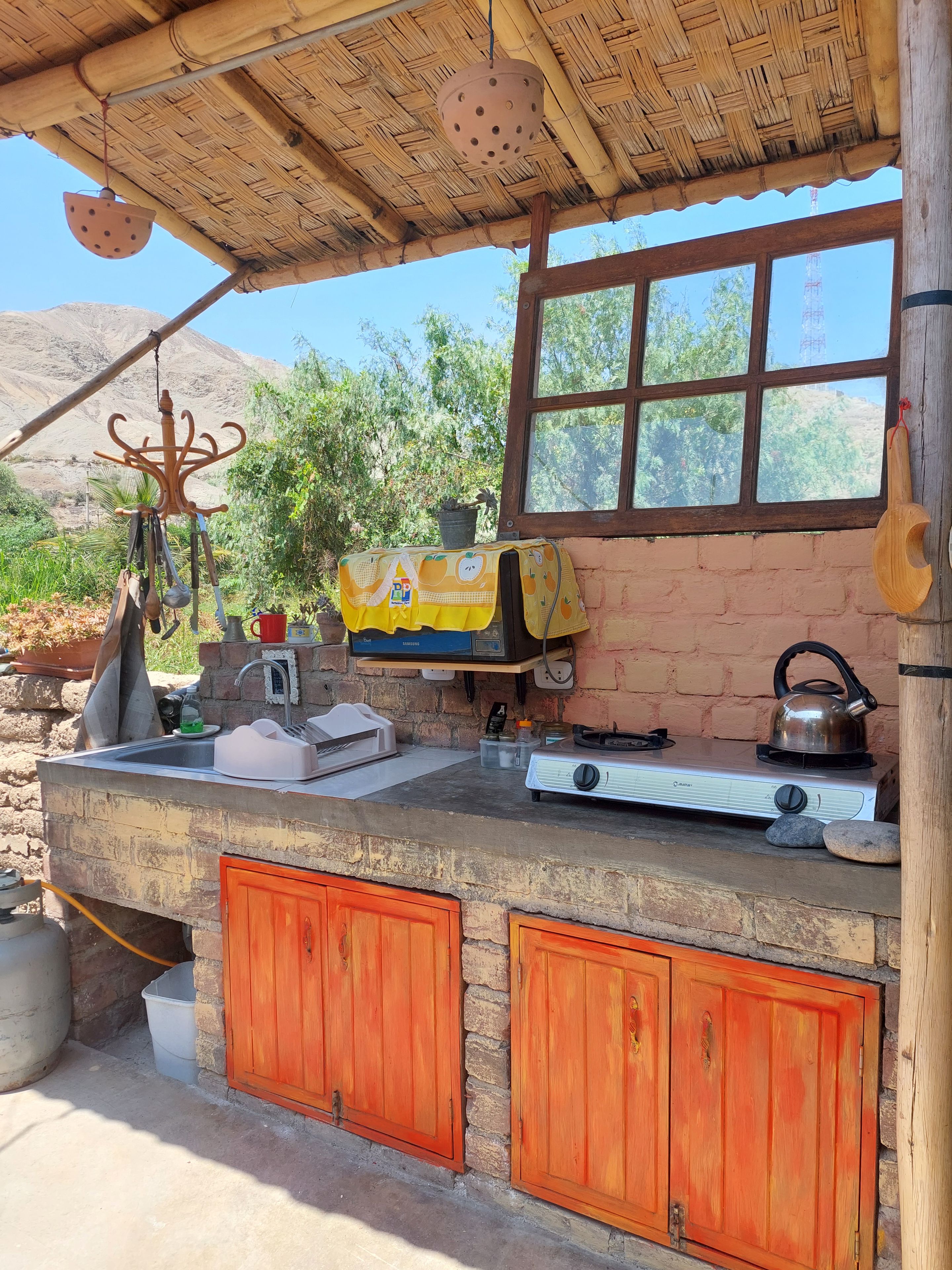 Cocina rústica al aire libre con mostrador de ladrillo, estufa a gas, tetera y colgador para ollas.