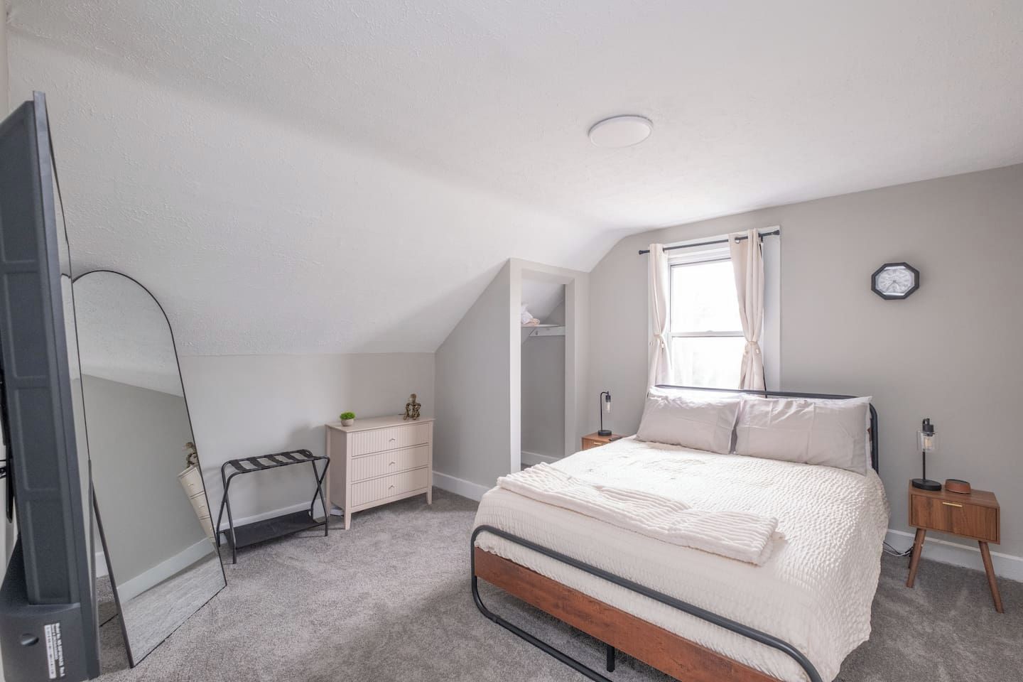 A bedroom with a bed, a dresser, a mirror, a window, and a clock on the wall.