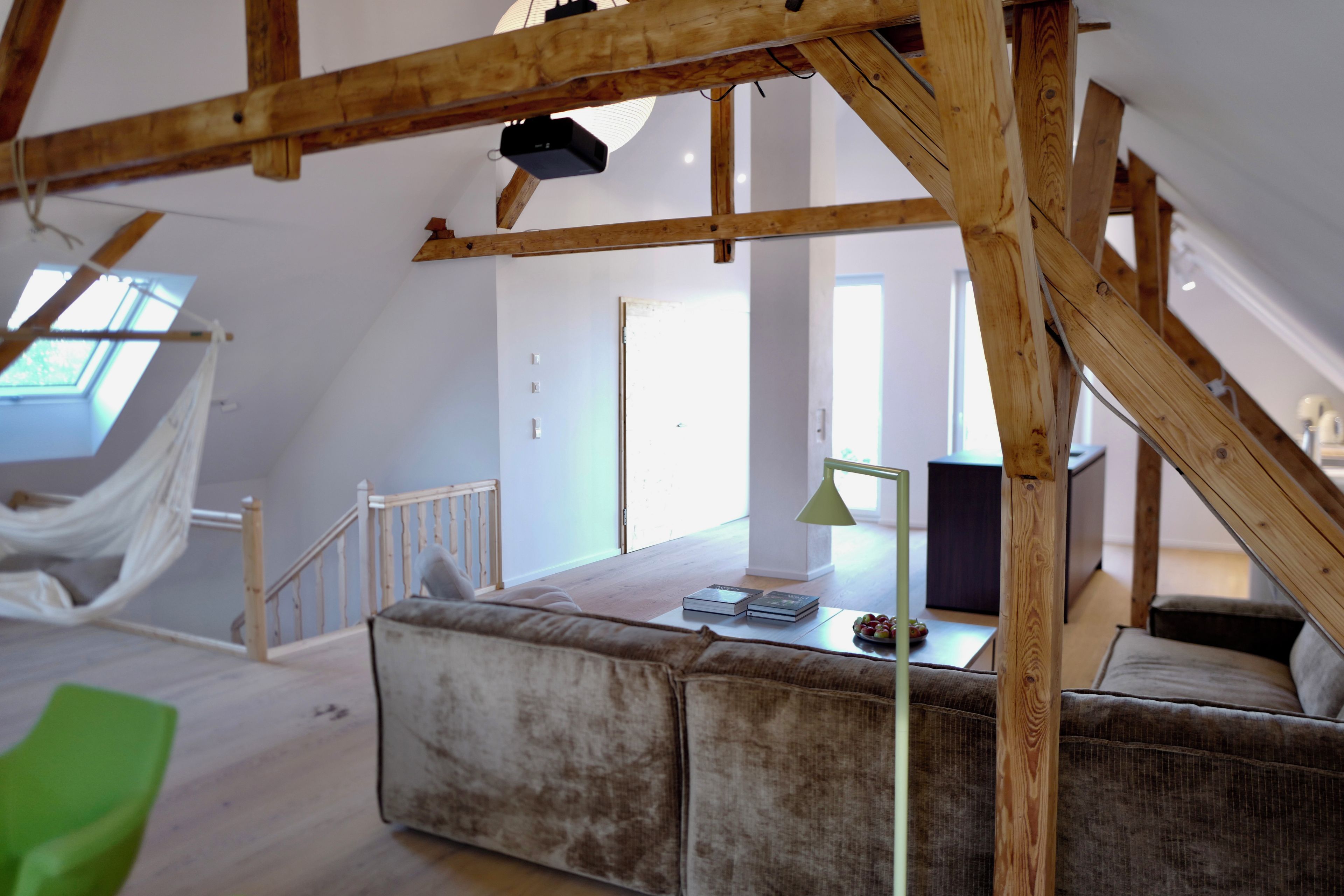 A loft-style room with exposed wooden beams, a green chair, a sofa, and a table with books and a lamp.