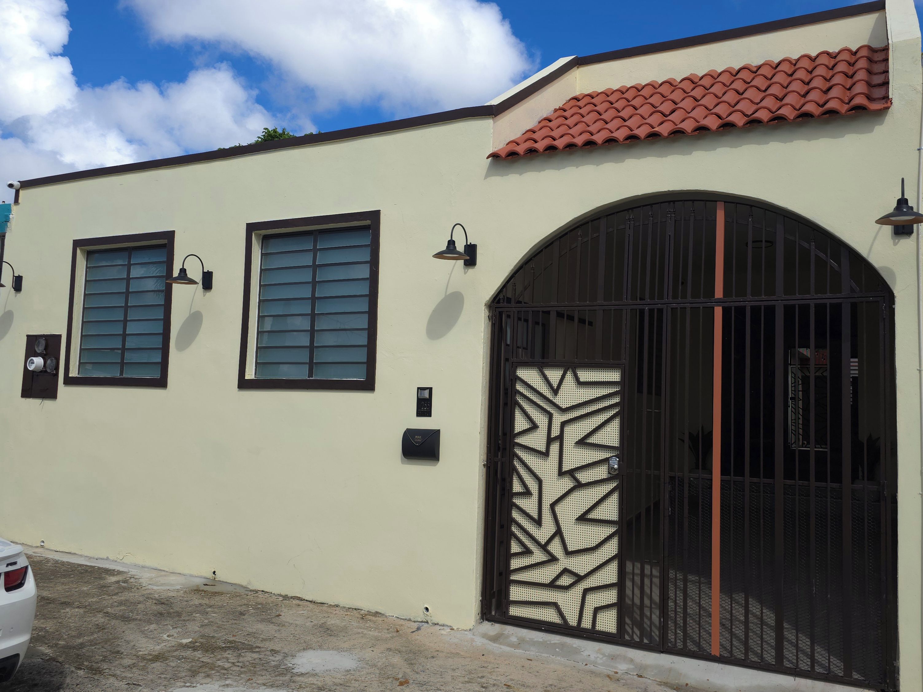 A building with a beige facade, two windows with shutters, a large arched entrance with a decorative metal gate, and a red-tiled roof.