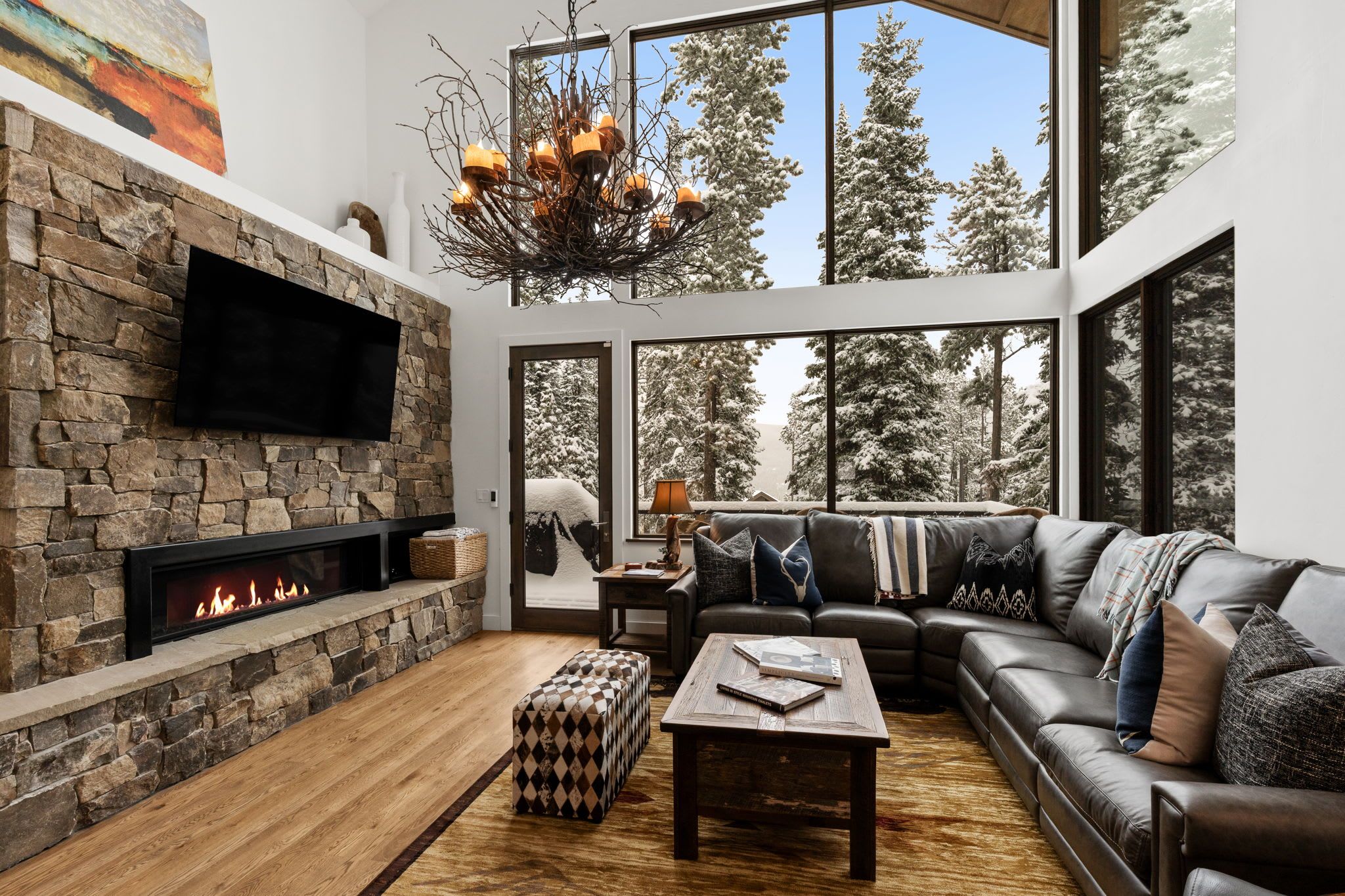 A modern living room with a stone fireplace, leather sofa, and large windows offering a view of snow-covered trees.