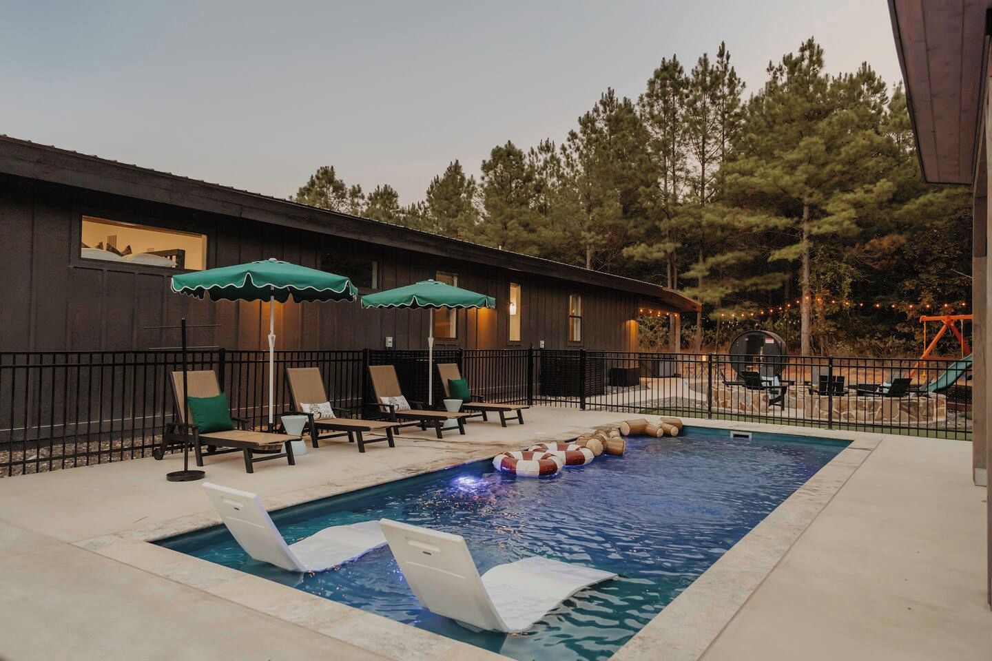 Una piscina con sillones de descanso y sombrillas, rodeada por una valla de madera y árboles.
