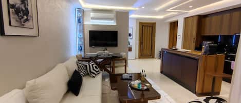 A modern living room with a sofa, coffee table, and a TV. There is a kitchen area with wooden cabinets and a dining table.