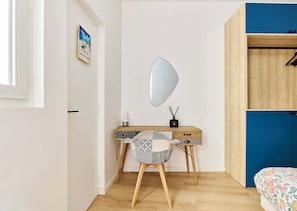 Quarto com escrivaninha de madeira, espelho, cama com colcha florida e guarda-roupa azul.